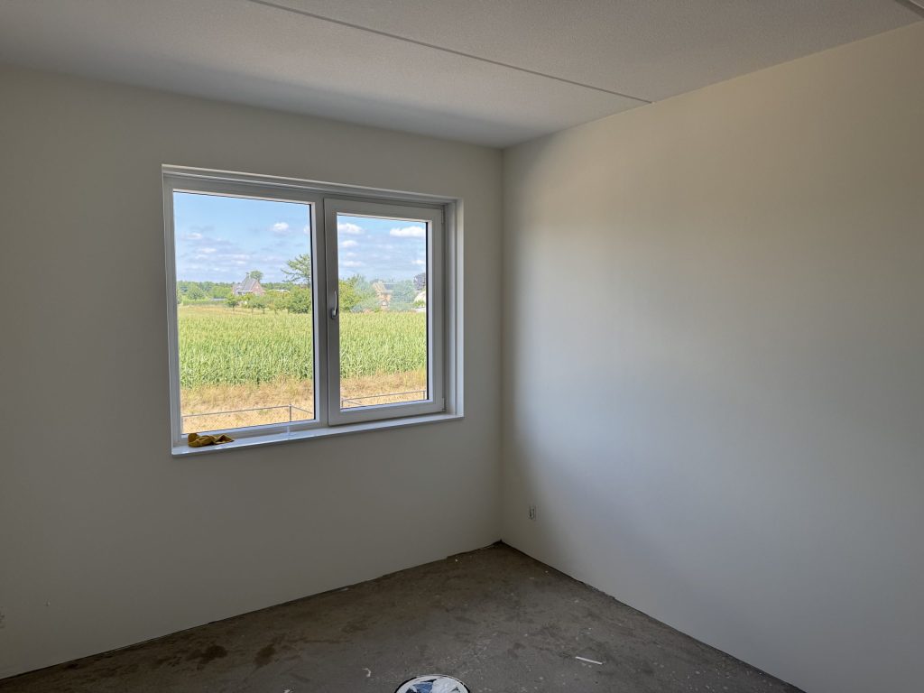 Slaapkamer met kale witte wanden en een raam dat uitkijkt op een maisveld